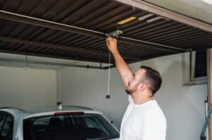 Man in a white shirt manually opens a garage door above a parked car in a garage.