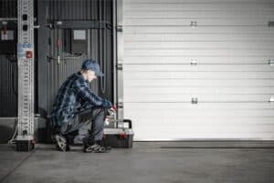 Person in a cap and plaid shirt kneels beside a toolbox in a garage, facing a closed white garage door.
