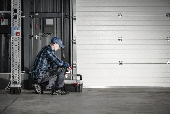 Person in a cap and plaid shirt kneels beside a toolbox in a garage, facing a closed white garage door.