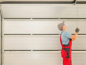 Worker in a red overall and gray cap manually adjusts a metal garage door with a pulley system inside a garage.