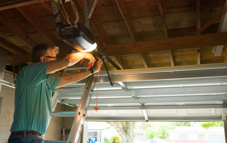 A person on a ladder is repairing a garage door opener in a garage with open double doors, contemplating the questions of how best to ensure its smooth operation.