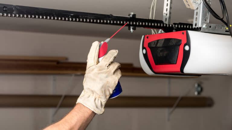 A gloved hand sprays lubricant on a garage door chain mechanism with a red and white spray can, raising questions about maintenance efficiency.