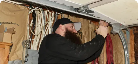 A person wearing a black cap is working on the overhead mechanism of a garage door, surrounded by wiring and insulation.
