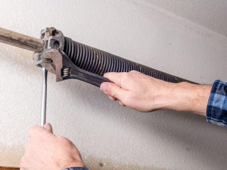 A person focuses on garage door repair, using a wrench to adjust the spring while carefully holding a metal rod to tighten the mechanism.