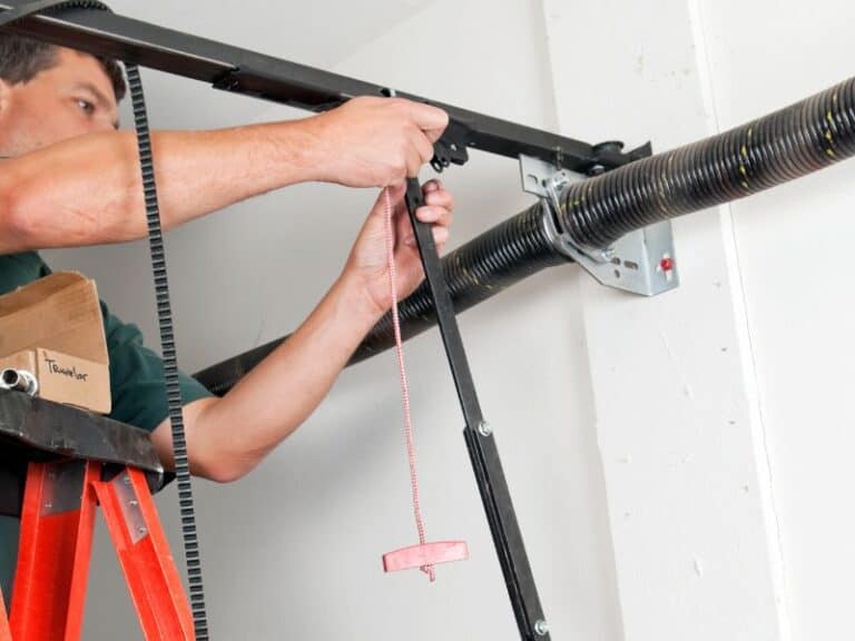 A person on a ladder is expertly performing a garage opener installation, skillfully adjusting the mechanism with black rails and a chain.