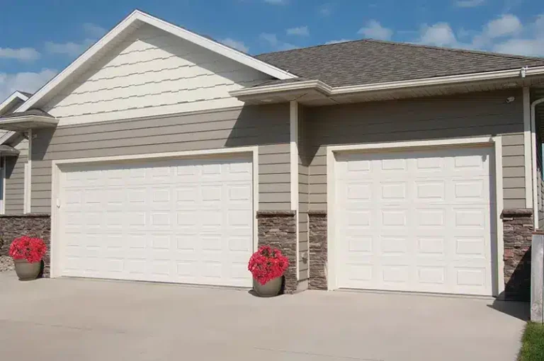 A beige double garage with pristine white doors stands side by side. Two potted red-flowered plants sit in front, under a clear blue sky, perfect for a day of garage door replacement.
