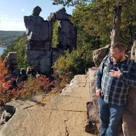 A person stands next to a rocky path with a view of a natural rock formation and trees, near a cliff edge—an escape from the usual office careers.