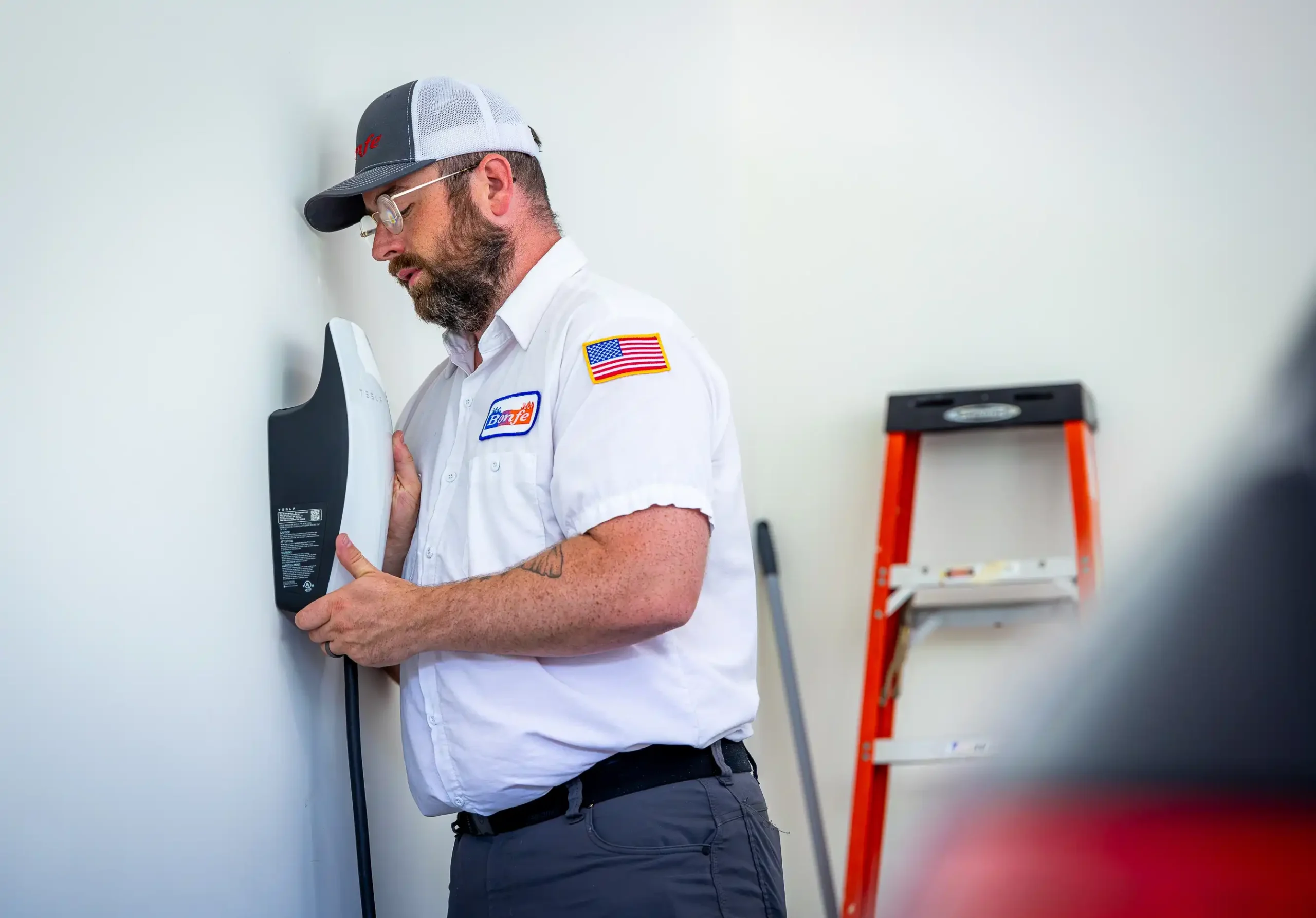 A technician in uniform completes a car charging install, mounting the device on the wall next to a ladder in a garage-like setting.