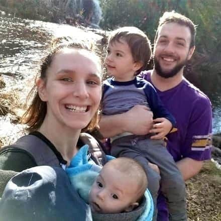 A smiling family of four by a creek, with two adults and two young children enjoying time away from their busy office careers. One adult holds a child, while the other carries a baby in a front carrier.