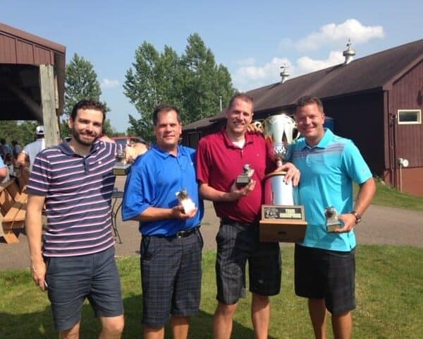 Four men, looking like they just nailed a big plumbing service technician challenge, stand outside holding small trophies. One also boasts a large trophy. They're in casual shirts and shorts, smiling with a barn and trees creating the perfect backdrop for their victorious moment.