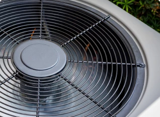 Close-up of an outdoor air conditioning unit with visible fan blades and a protective grille, set against a background of green foliage.