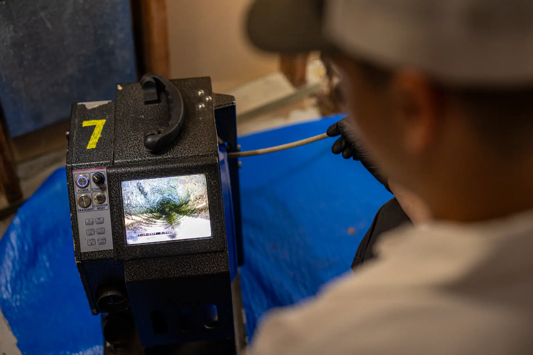 A person operates a pipe inspection camera, viewing a live feed of the inside of a pipe on a monitor, positioned on a blue tarp, as part of an efficient drain cleaning process.