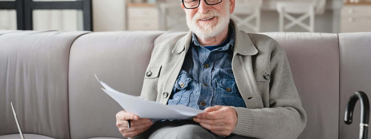 Grandfather looking at camera checking pension documents, loan, e-banking, paperwork with laptop
