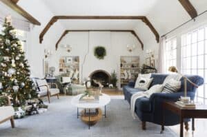 A bright living room with exposed wooden beams, a decorated Christmas tree, a blue sofa, bookshelves, and a fireplace adorned with a wreath creates a cozy winter wonderland atmosphere.