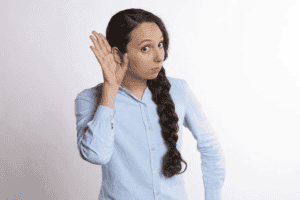 Woman with long dark braid in a light blue shirt holding her hand to her ear, appearing to listen closely, as if curious about when to replace a boiler, against a plain light background.