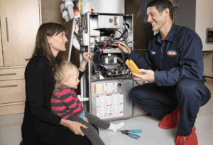 A repair technician in uniform services a furnace while a woman and child watch. Tools are on the floor nearby.