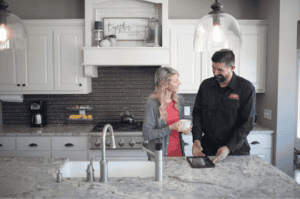 A woman holding a mug talks with a male technician in uniform, who is using a tablet, in a modern kitchen with white cabinets and granite countertops.