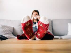 A person in St. Paul Minneapolis relaxes on a sofa with feet up on a table, wearing red socks with reindeer and snowflake patterns, and sipping from a white mug—enjoying warmth thanks to a high efficiency furnace.