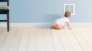 A baby in a white onesie crawls on a light wooden floor toward a wall vent in a minimally furnished Minnesota home, highlighting the importance of indoor air quality.