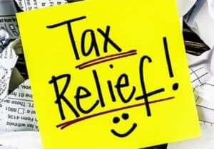 A yellow sticky note reading "Tax Relief!" with a smiley face sits atop a pile of assorted papers, hinting at possible savings like water heating tax credits recently discussed in Congress.