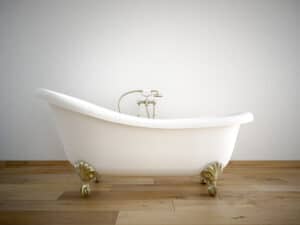 A vintage-style white clawfoot bathtub with gold feet sits on a wooden floor against a plain white wall—perfectly paired with the best water heaters from Hero Plumbing for a luxurious soak.