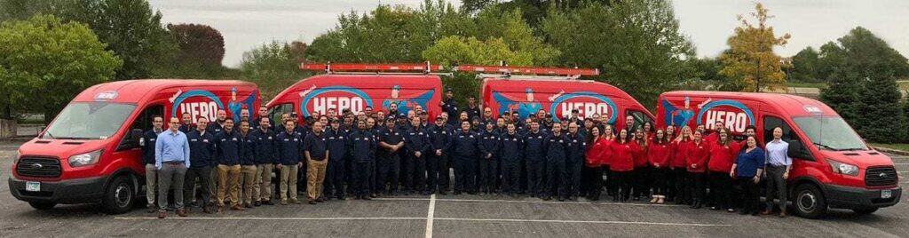A large group of people pose in front of three red HERO-branded vans in a parking lot, ready to answer your Request Service needs, with trees and greenery adding to the background.