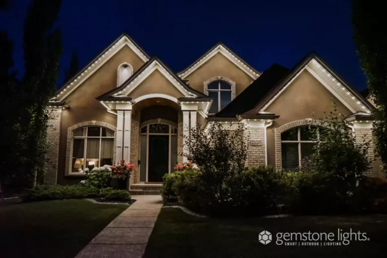 A house with peaked rooflines is illuminated by exterior trim lights at night, featuring a front porch, columns, and landscaped yard.