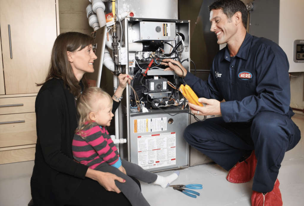 A technician in uniform repairs a furnace while a woman and a young child watch nearby in a basement or utility room.
