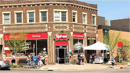 A brick corner building houses Uptown shop, with red awnings and people standing outside near a white tent and a bike rack on a sunny day.