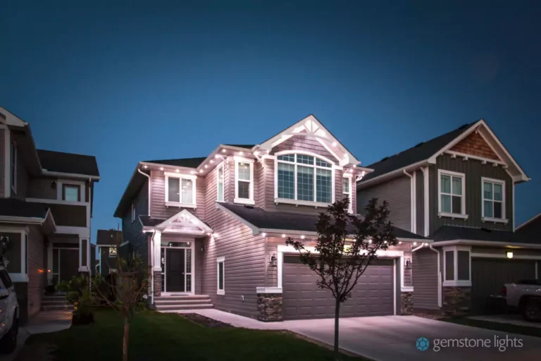 Two-story suburban house with gray siding and white trim, illuminated by accent lighting along the roofline at dusk.