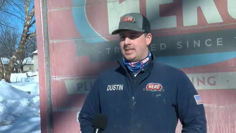 A man wearing a "HERO" branded hat and jacket labeled "Dustin" stands outside in snowy conditions, with a large "HERO" sign behind him and a microphone visible in the foreground.