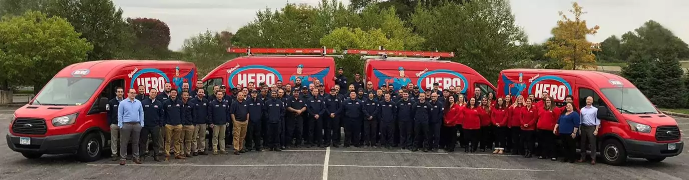 A large group of people stands in front of three red HERO-branded vans in a parking lot, with trees and greenery in the background.
