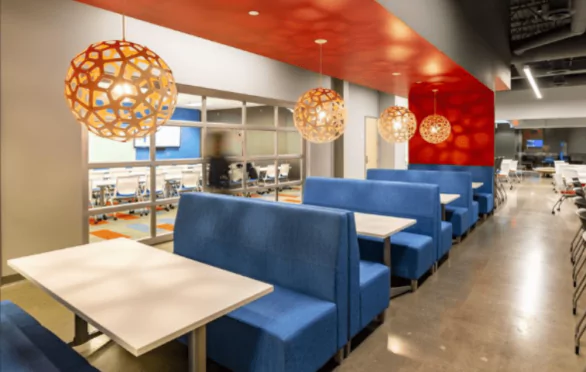 Modern office lounge area with blue booth seating and white tables, illuminated by geometric pendant lights, features a red accent wall and glass window overlooking a workspace—a stylish setting for Hero news updates in the Twin Cities.