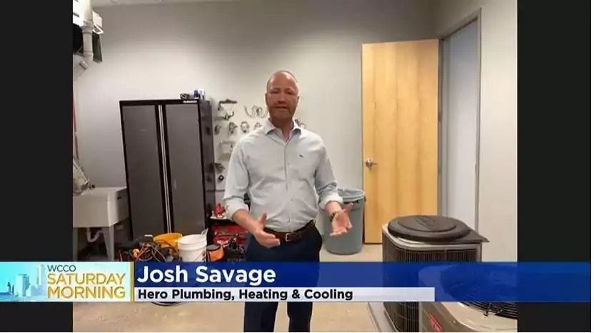A man stands in an indoor room with HVAC equipment and tools. On-screen text reads: "Josh Savage, Hero Plumbing, Heating & Cooling" and "WCCO Saturday Morning.