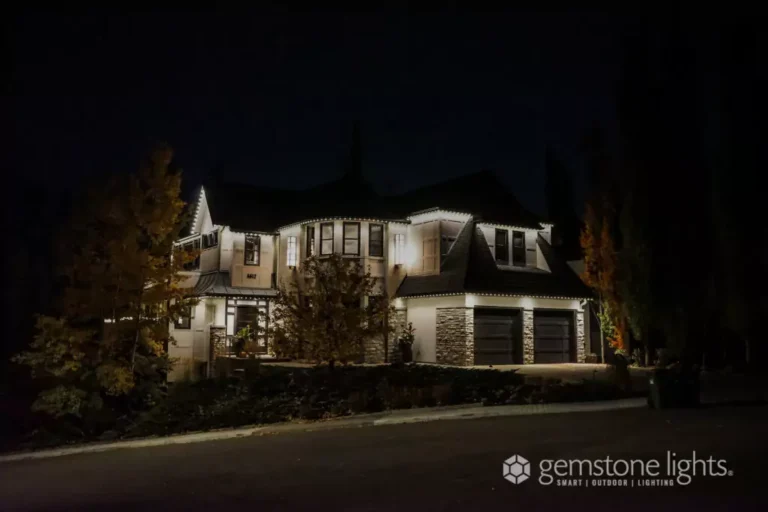 A two-story house at night with its rooflines and garage outlined by white LED lights. Trees partially frame the home, and the Gemstone Lights logo is visible in the corner.