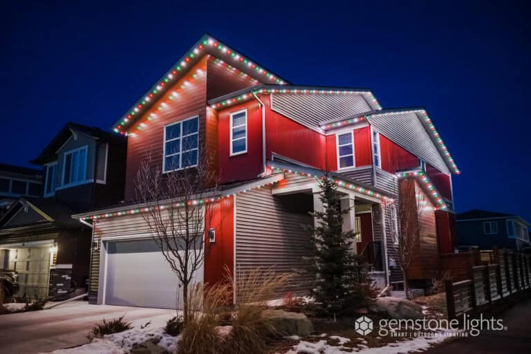 Two-story red and gray house at night with multi-colored lights outlining the roof; patches of snow are visible on the ground.