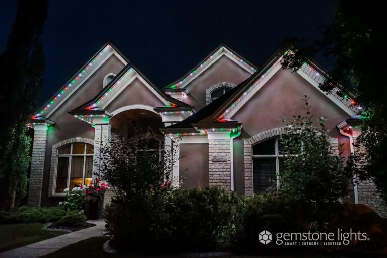 A house at night with multi-colored smart outdoor lights outlining the roof and eaves; "gemstone lights" branding appears at the bottom right.