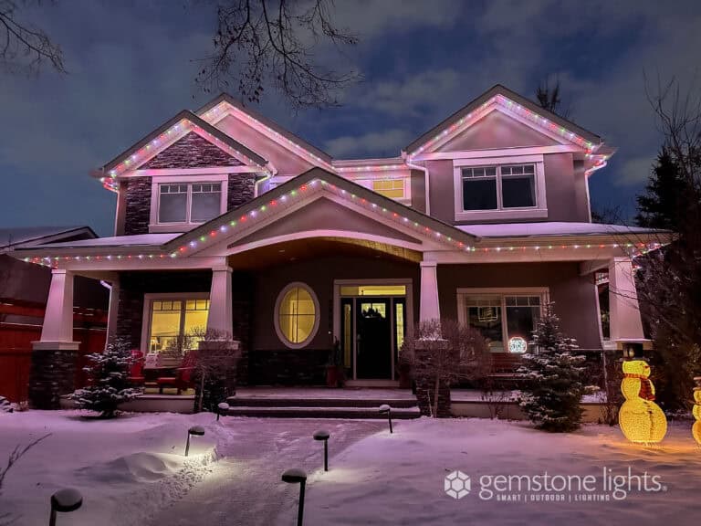 A two-story house with snow-covered yard is decorated with multicolored LED lights along the roofline and an illuminated snowman in front.