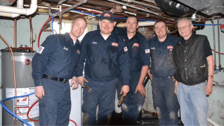 Five men stand in a basement near a water heater; four wear matching work uniforms, representing Hero giving back to Twin Cities homes, while one is dressed casually. Pipes and equipment are visible in the background.