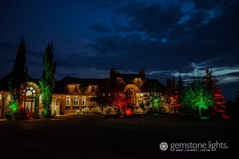 A large house is illuminated at night with red and green exterior lights, highlighting trees and architectural features. The sky is dark and cloudy.