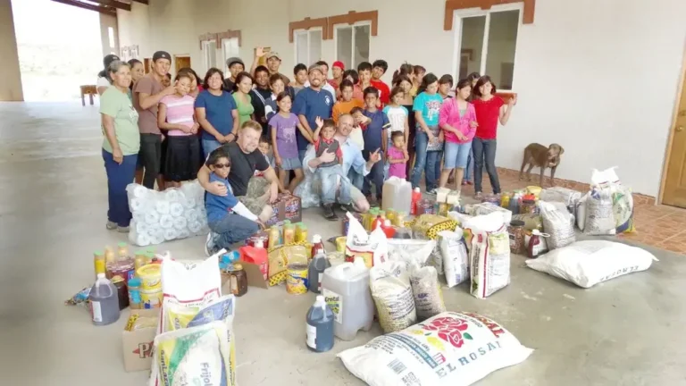 A large group of people, including children and adults, pose indoors with various food supplies and groceries arranged on the floor in front of them—showcasing a true hero giving back to the Twin Cities. A dog stands near the back.