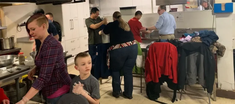 Several adults and a child work together in a crowded kitchen, preparing food and cleaning—true heroes giving back to the Twin Cities community, with coats draped over a chair in the background.