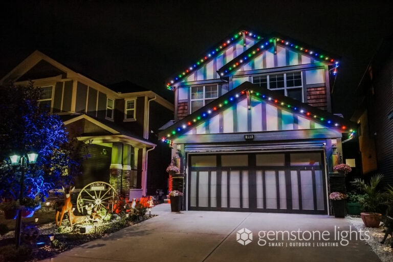 Two-story house at night with multi-colored decorative lights outlining the roof and garage; neighboring house and front yard decorations are also visible.