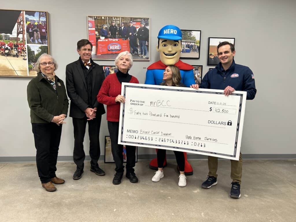 Five people pose indoors with a $43,500 donation check to the Minnesota Breast Cancer Coalition, joined by someone in a HERO mascot costume from Hero Home Services.