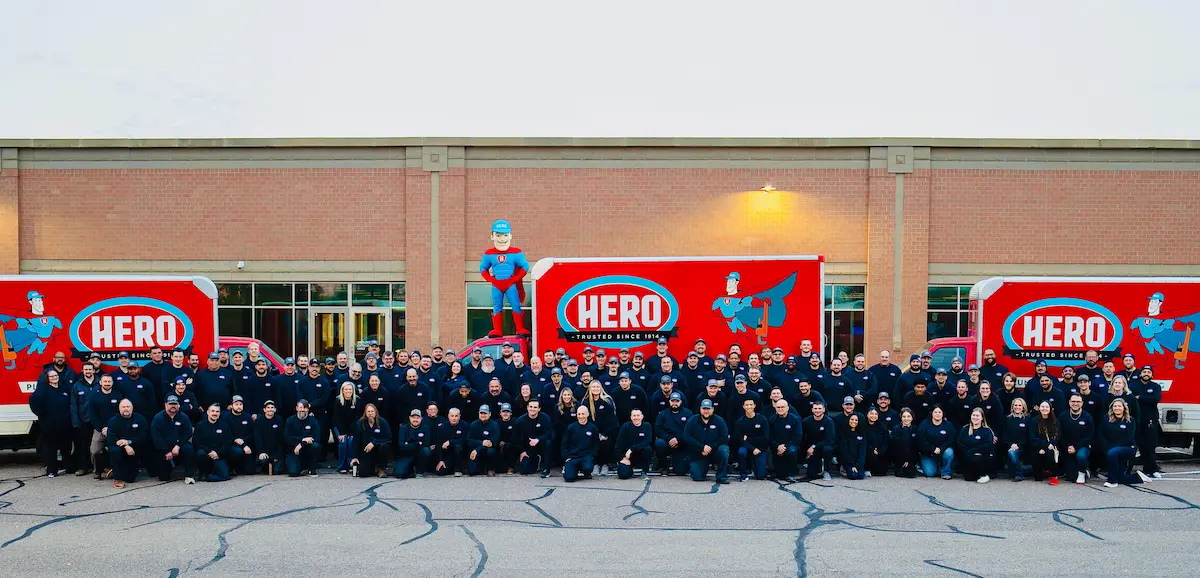 A large group in matching dark outfits poses in front of two red Hero-branded trucks and a brick building, highlighting Hero community service in the Twin Cities, with a superhero statue above the trucks.