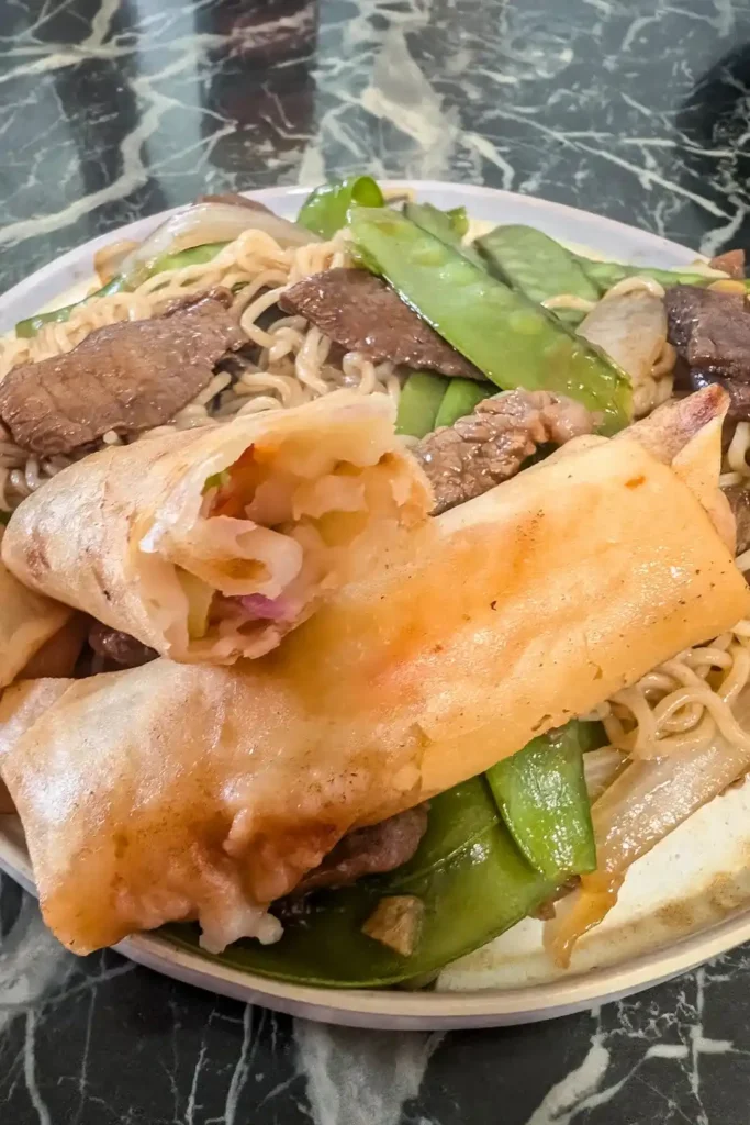 inside a shrimp spring roll that rests on another on a plate of beef and snow pea ramen noodles