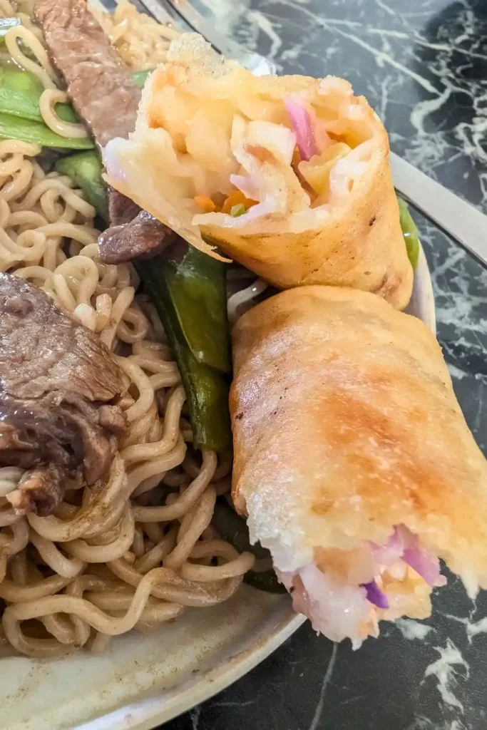a spring roll broken open on the side of a plate with beef noodles on black marble table