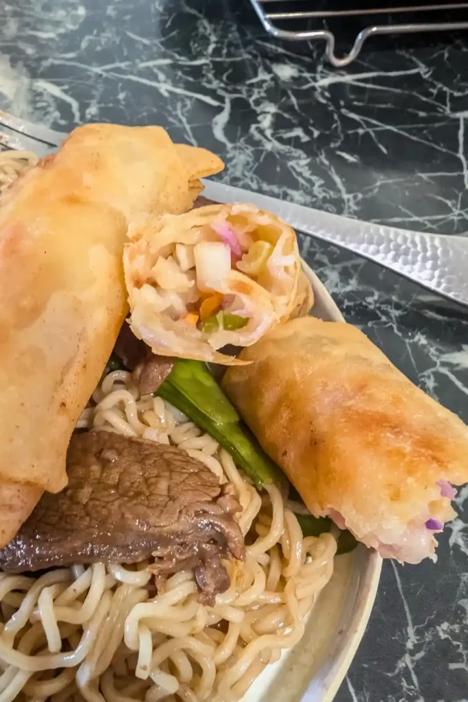 a fork resting in background on the plate with beef noodles and spring rolls. One is broken in half the other is whole