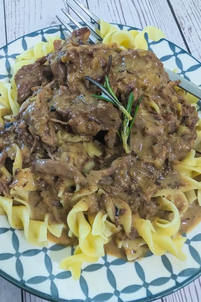 a blue and white plate with the beef and noodles topped with a sprig of rosemary a fork resting at the side