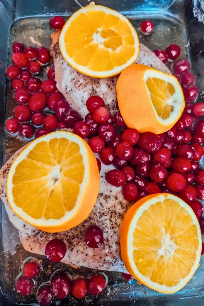partially cooked turkey breast with cranberries and orange slices in glass baking dish